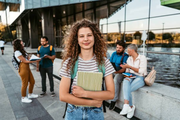 Portrait of a beautiful smiling university student standing on campus with friends in the background.